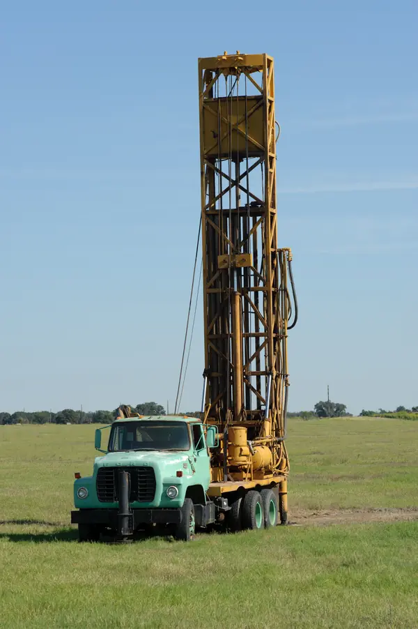 A borehole drilling rig set up on a grassy plot, its mast raised over the hole